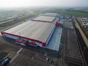 Aerial view of a large TIA, Lancaster Warehouse with a red and white exterior, adjacent parking lot, and surrounding green fields.