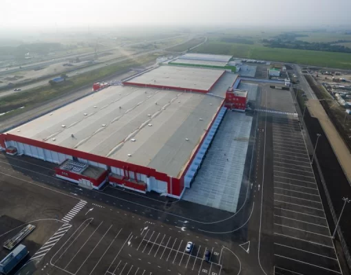 Aerial view of a large TIA, Lancaster Warehouse with a red and white exterior, adjacent parking lot, and surrounding green fields.